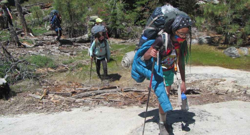 Students wearing backpacks make their way across a grassy and rock landscape. They brace themselves with hiking poles.